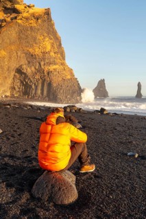 A man in an orange jacket sits on a rock on a beach. The sky is blue and the sun is shining. The