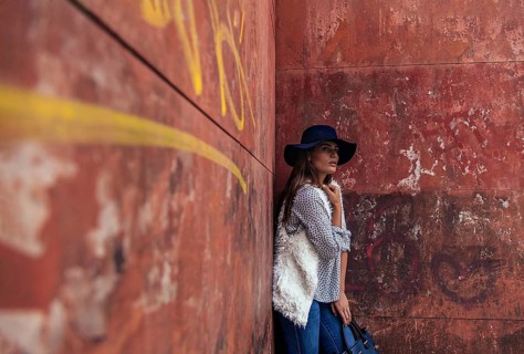 A fashionable woman in a blue hat leans against a weathered red wall adorned with graffiti. She