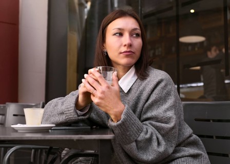 A thoughtful woman in a gray sweater holds a glass while sitting at an outdoor bar table. The cozy