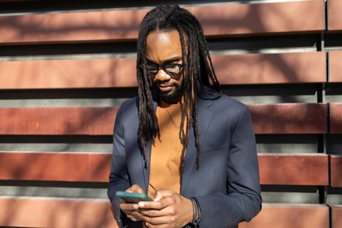 A cuban business professional with glasses and dreadlocks stands outdoors in sunlight, using a