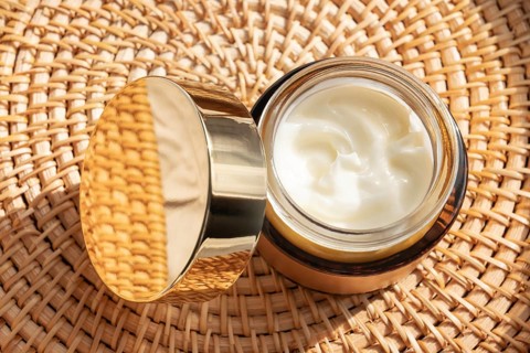 A luxurious skincare cream cosmetic with a golden lid rests on a woven mat. The rich cream inside