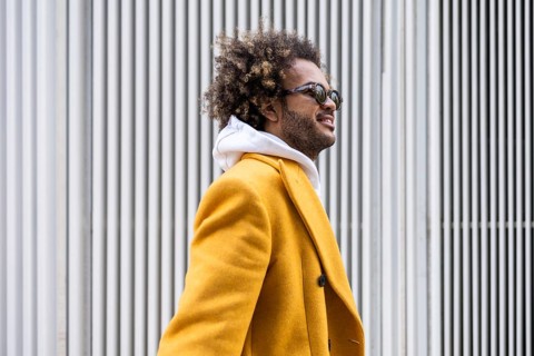 Afro man wearing a stylish mustard coat gazes confidently to the side. His sunglasses reflect a