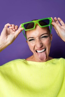A vibrant middle aged woman with a shaved head wearing bright green sunglasses and a yellow sweater