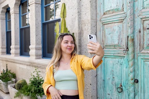 A woman takes a selfie on a charming street, wearing sunglasses and a yellow shirt. She stands in