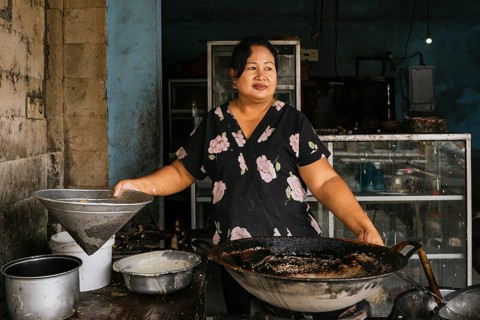 A woman stands in a rustic kitchen, preparing traditional dishes with a large wok. The worn walls