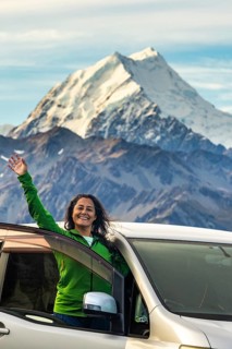 A woman stands joyfully in front of her car, surrounded by the breathtaking autumnal beauty of