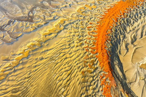 Aerial image showcasing striking orange and yellow sand patterns resembling waves on a beach,