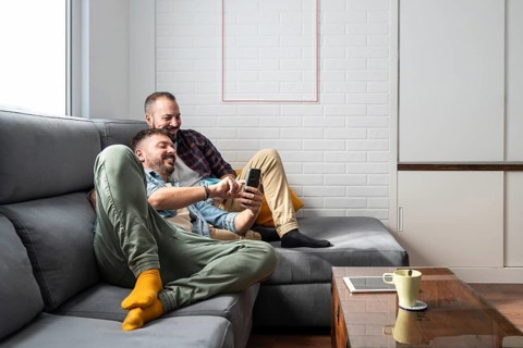 A happy gay couple relaxes on a cozy couch, smiling as they look at a smartphone. The setting is a