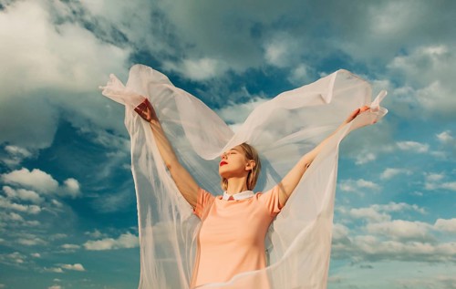 A woman with outstretched arms embraces the breeze, her sheer fabric flowing gracefully. A serene