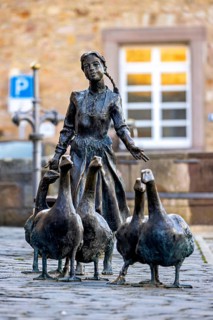 Bronze statue of a girl with geese on cobblestones, in the background a building with white