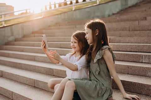 Two young girls sit on sunlit steps in summer, happily taking a selfie. They are enjoying a sunny