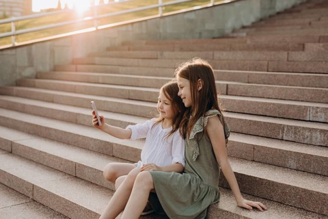 Two young girls sit together on sunlit stone stairs, smiling as they take a selfie. The warm glow