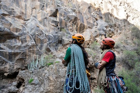 Two climbers equipped with helmets and ropes study a rugged cliff, preparing for a traditional