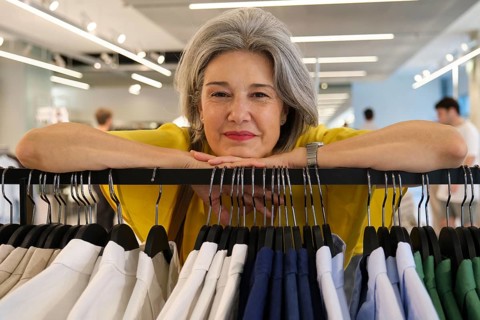 A mature woman with gray hair and a bright yellow shirt smiles warmly while leaning on a clothing