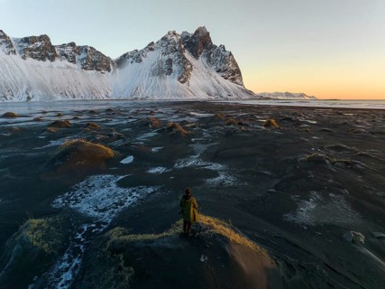A man stands on a textured, frost-covered dune, gazing at the towering, snow-draped mountains of