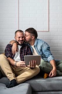 A loving gay couple sits on the couch, sharing a sweet moment while using a tablet. They embrace