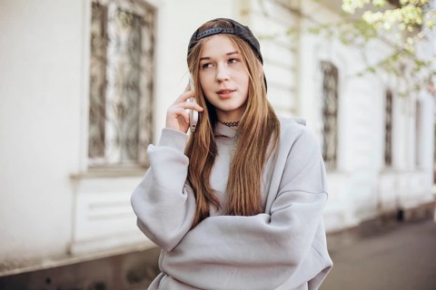A teenage girl wearing a casual hoodie and cap is engaged in a call on her smartphone outside. She