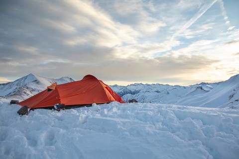 A vibrant orange tent sits atop a snow-covered peak, surrounded by majestic mountain views under a