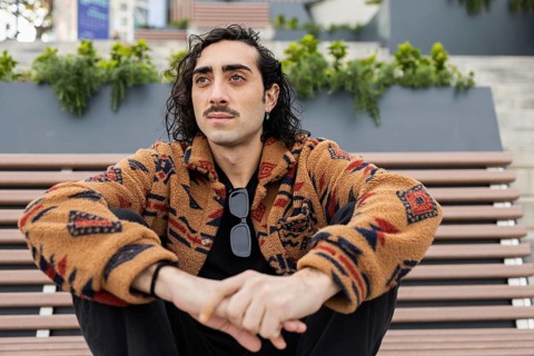 A man with curly hair and a mustache sits on an outdoor bench wearing a patterned jacket and