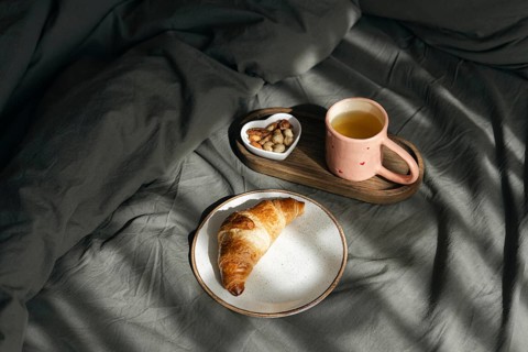 A golden homemade croissant rests on a white plate, accompanied by a wooden tray holding a pink cup