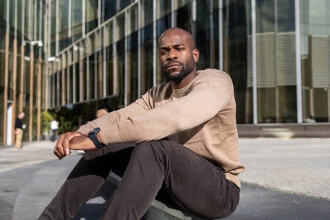 A cuban man in casual attire sits outside a modern glass building, embodying a relaxed urban vibe.