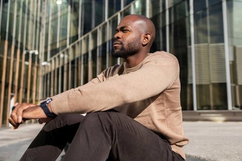 A cuban man in a casual outfit sits outdoors, leaning against a modern glass building. He appears