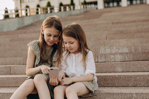 Two young girls sit together on outdoor steps in summer, engrossed in a smartphone. They share a