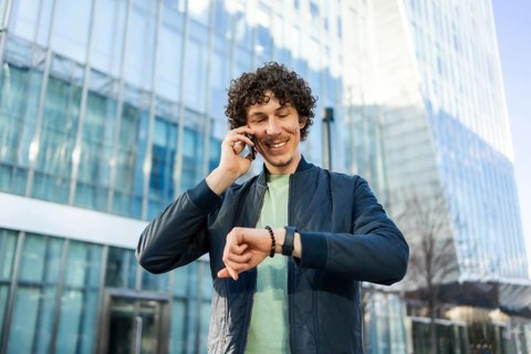 Smiling man in casual attire checks the time on his smartwatch while talking on a smartphone.