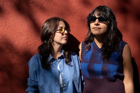 A multiethnic lesbian couple shares a moment outdoors, standing against a sunlit red wall. Both