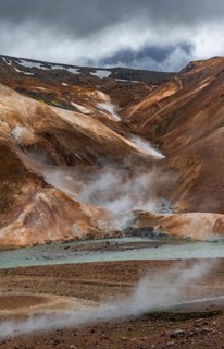 Steaming stream between colourful rhyolite mountains with snowfields, Hveradalir geothermal area,