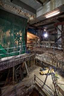 An interior construction site with exposed beams, scaffolding, and safety nets. Illuminated by