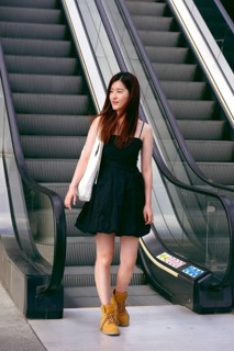 A young asian woman in a black dress stands confidently on an escalator. Her brown hair is loose,