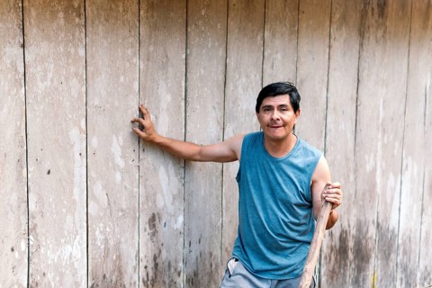 A middle-aged man stands confidently in the Colombian countryside, leaning against a rustic wooden