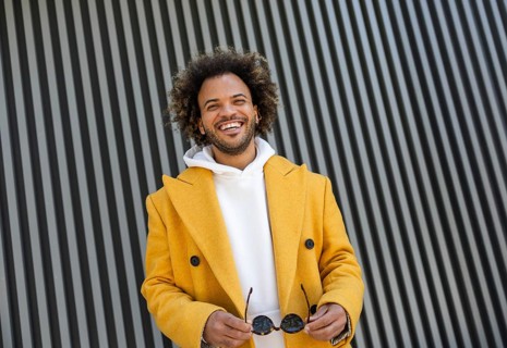 A cheerful Afro man in a yellow coat holds sunglasses, standing in front of a modern striped