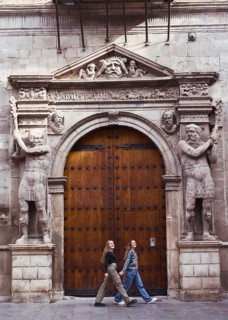 Two young woman friends casually walk past an ornate wooden door framed by intricate stone carvings