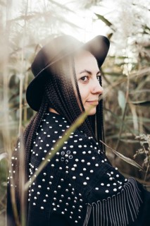 A woman in a unique studded jacket and black hat stands amidst tall grasses at sunset, exuding a