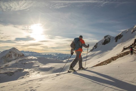 Two backcountry skiers ski down a pristine snowy mountain under a bright sky. Equipped with