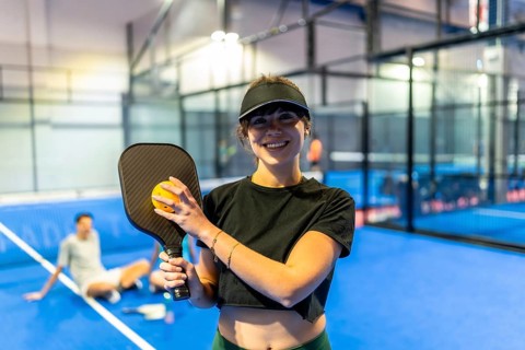 Smiling young woman holding a pickleball paddle and dimpled plastic ball, standing on a blue court