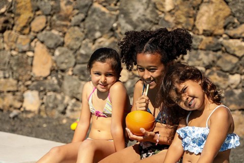Three children in swimsuits sit happily against a stone wall. They enjoy refreshing drinks, basking