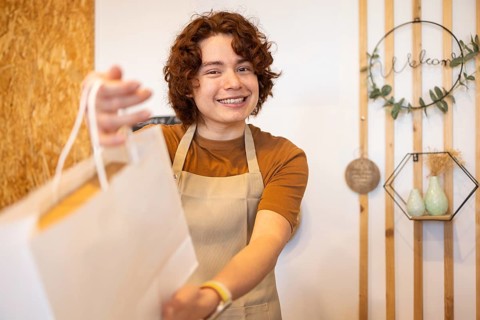 A cheerful shop assistant in an apron hands a paper bag to a customer, creating a warm and