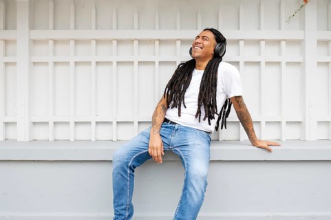 A joyful man with long dreadlocks and tattoos relaxes against a white fence, wearing headphones and