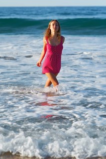 A woman in a vibrant pink polka dot dress joyfully runs through the surf at a sunlit beach in