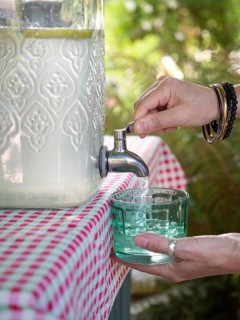 Fresh lemonade in large glass bottle with tap. Hands serving in glass. Refreshing summer drink.