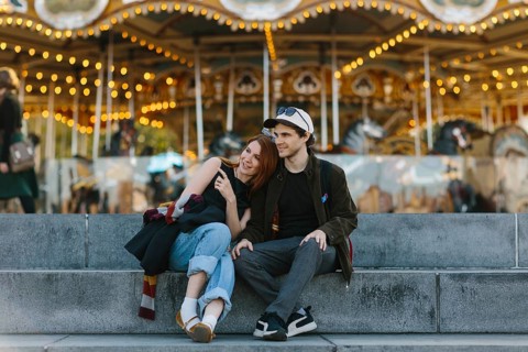 A couple sits on steps, embracing in front of a lit up carousel in Brooklyn Bridge Park. The joyful