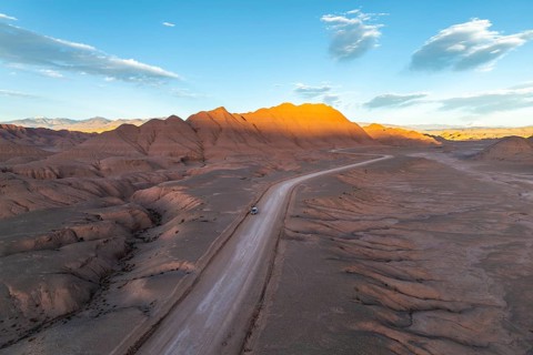 Aerial view of a vehicle in a empty desert road winding through rugged, sunlit mountains at dawn in