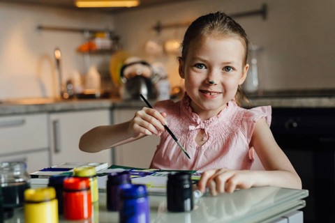 A joyful child with a painted nose engages in a creative painting activity at home. She holds a