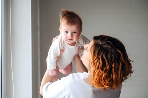 A mother lovingly holds her smiling newborn baby girl near a sunlit window, capturing a moment of