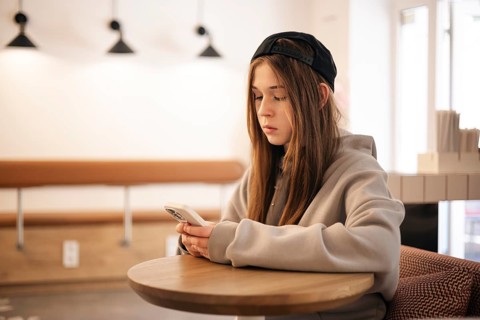 Teenage girl sitting at a cafe table using her smartphone. She is wearing a casual hoodie and cap,