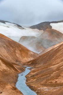 Steaming stream between colourful rhyolite mountains with snowfields, Hveradalir geothermal area,