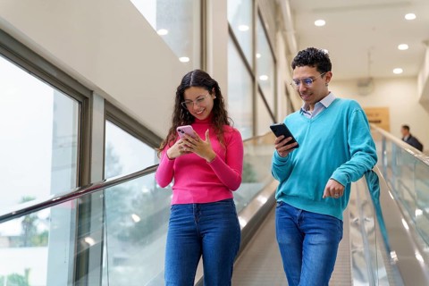 Young man and woman, Gen Z friends, enjoy using their smartphones while riding an escalator in a